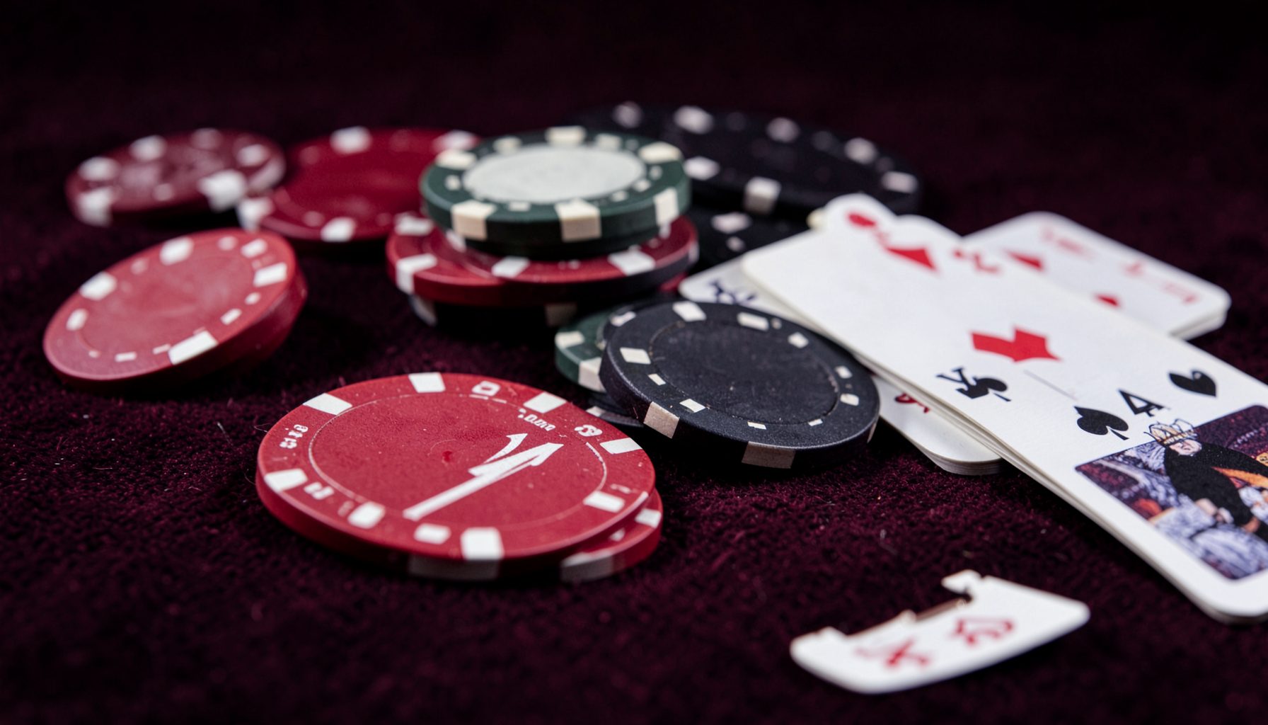 A close-up of poker chips and playing cards on a dark felt table, rich crimson and black tones