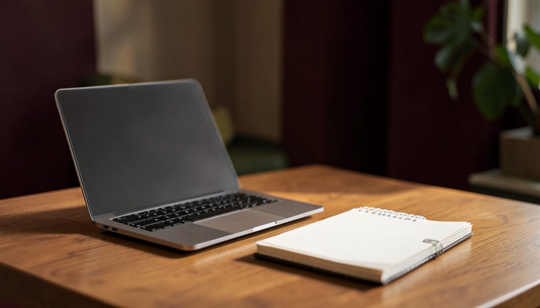 A warm, softly lit workspace with a laptop and notebook on a wooden desk, deep maroon and olive accents in the background, calm and professional atmosphere